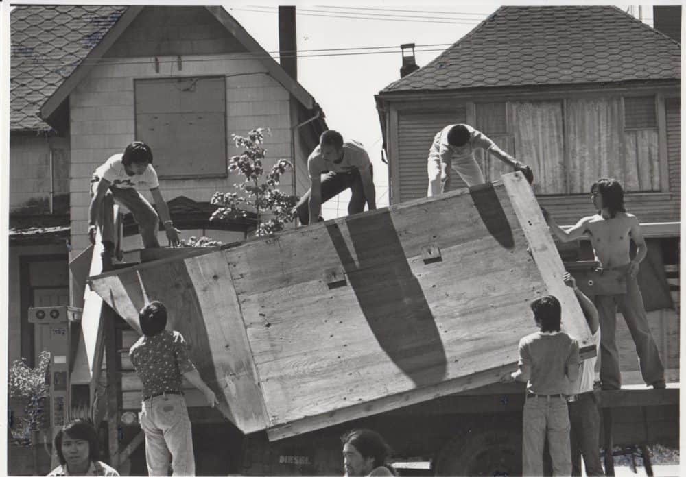 A black-and-white photo, taken in 1977, of five people taking a wooden structure off of the back of a truck.