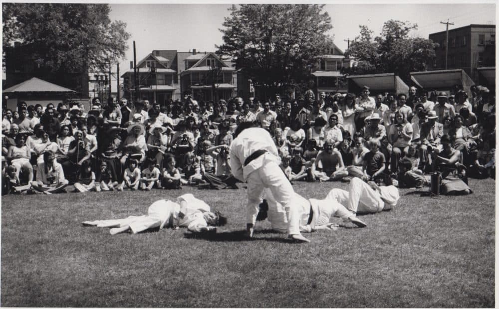 Black and white photo taken in 1977 of an outdoor martial arts tournament or demonstration. One person, with their back to the camera, stands over three others lying on the ground. All are wearing martial arts gi and black belts.